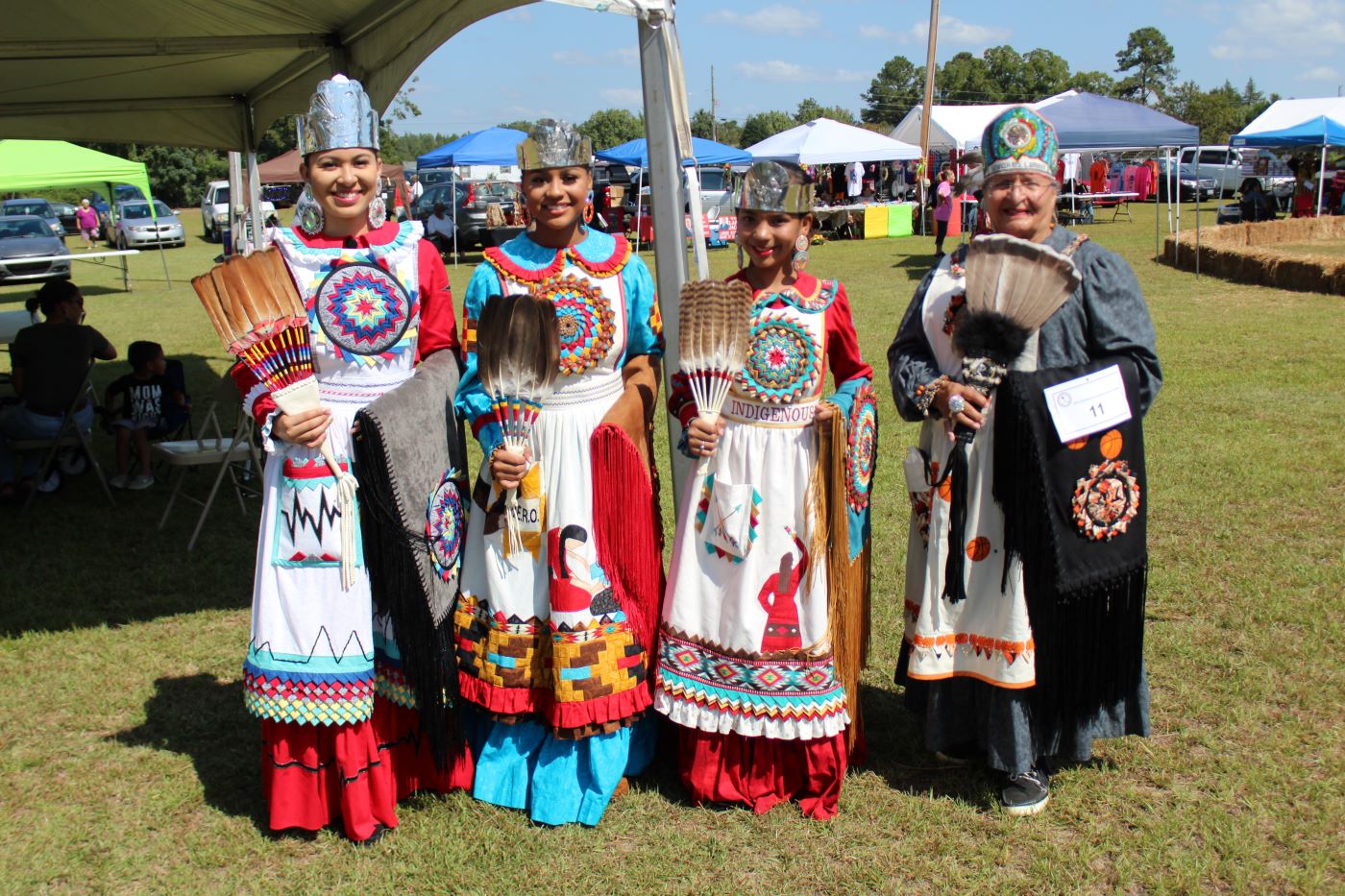 Four women of multiple generations are dressed in traditional native aprons and  how feather fans at the Lumbee Trib Homecoming in Lumberton, North Carolina. 