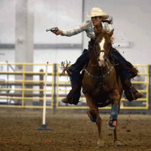 A woman riding a horse holding and shooting a revolver at a nearby target