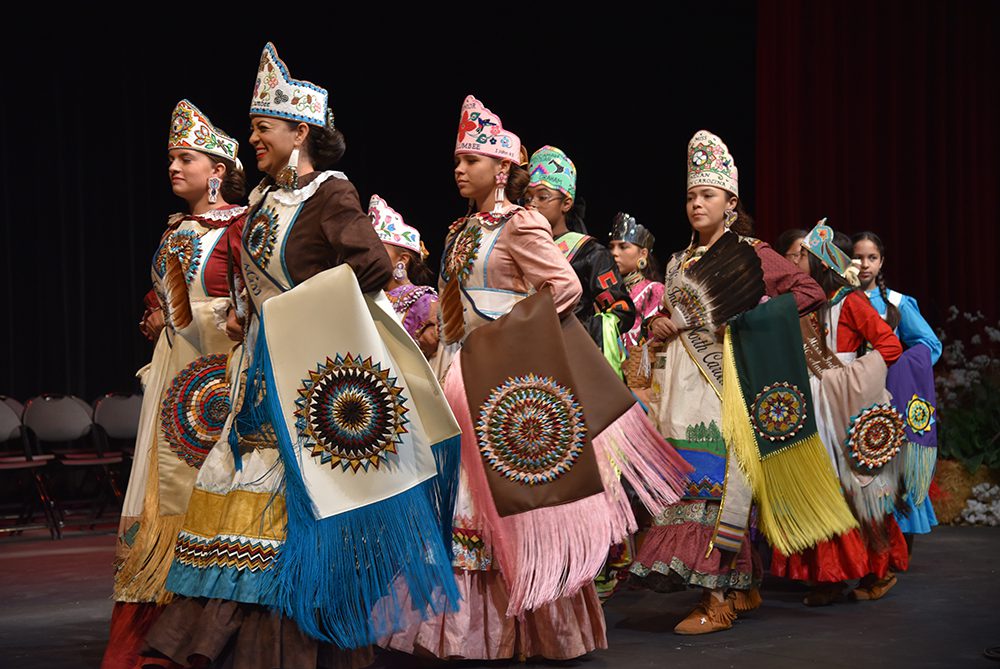 A group of Lumbee youth dressed in traditional regalia walk across a stage at the Givens Performing Arts Center.