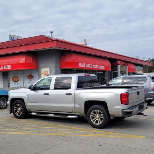 Exterior corner shot of Italy Grill Pizza & Subs in Lumberton, NC with lots of large trucks & SUVs in the parking lot