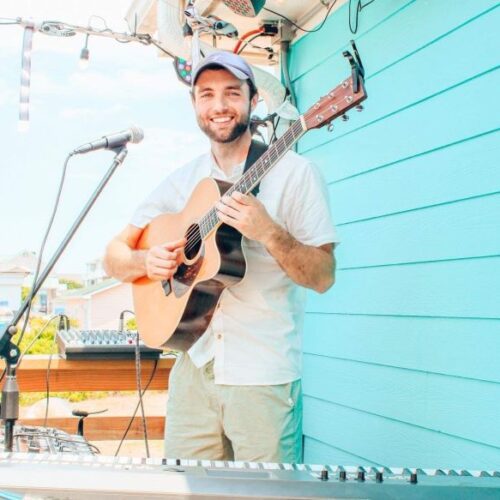 Matt Letch plays acoustic guitar on an outdoor deck in Lumberton, NC in front of a bright teal wall