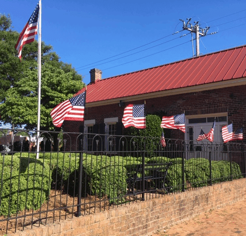 Exterior of a brick building with a red roof with a black metal fence topped with many American flags
