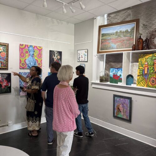 An older woman gazing upon a wall of assorted art pieces with a group of people behind her point at a different art piece.