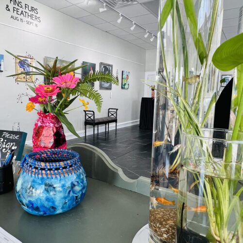 Water and plant filled vases on a desk with art hanging on a wall in the background at the Robeson County Arts Council.