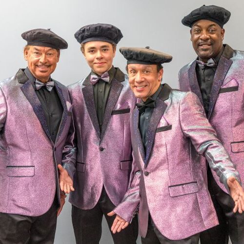 The Tams, wearing shimmering purple blazers, bow ties, and black berets, smile and pose together.