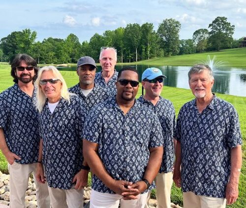 The Sand Band in matching patterned shirts pose outdoors near a pond and green hills
